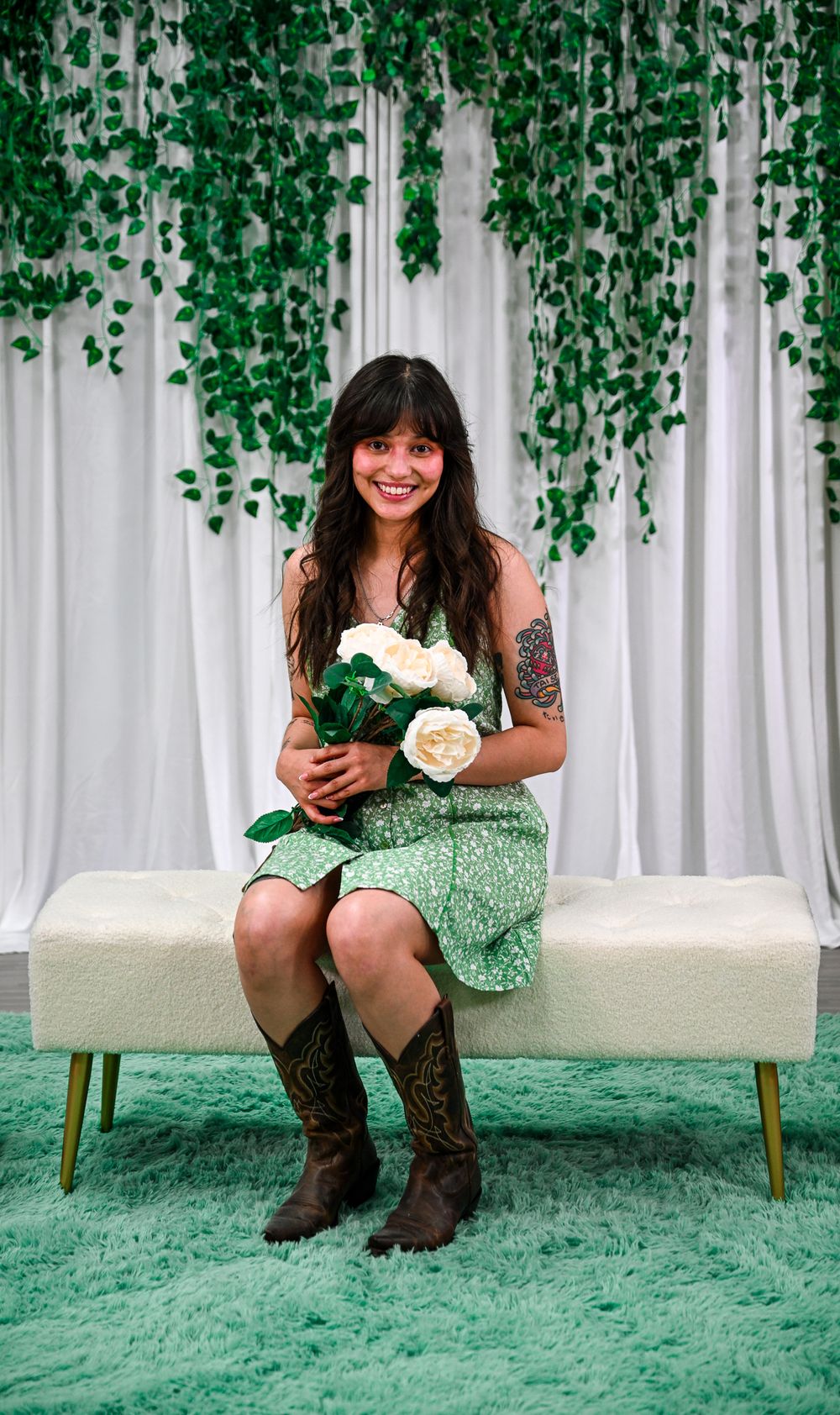 Smiling woman in green floral dress and cowboy boots sits on bench holding white roses with ivy backdrop.