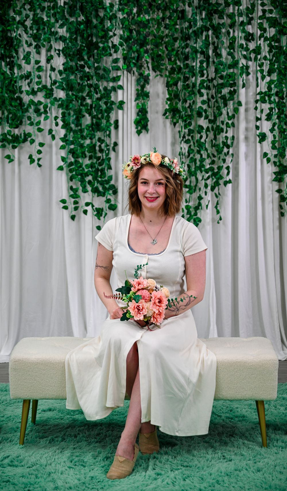 Woman in white dress with floral crown holds pink bouquet, seated on bench with green ivy backdrop.