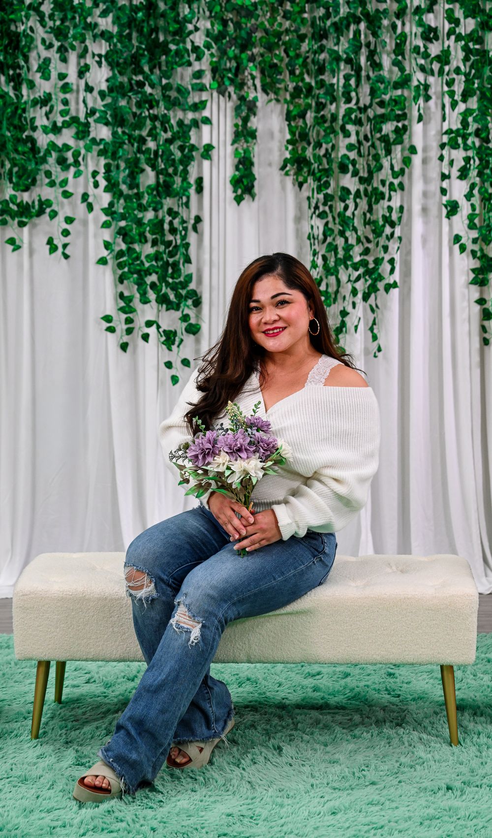 Woman in white sweater and jeans holds purple bouquet, seated on bench with green ivy backdrop.