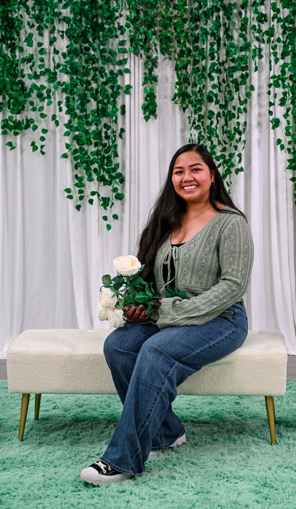 Young woman in sage green cardigan sits on white bench holding white roses, smiling before green ivy backdrop.