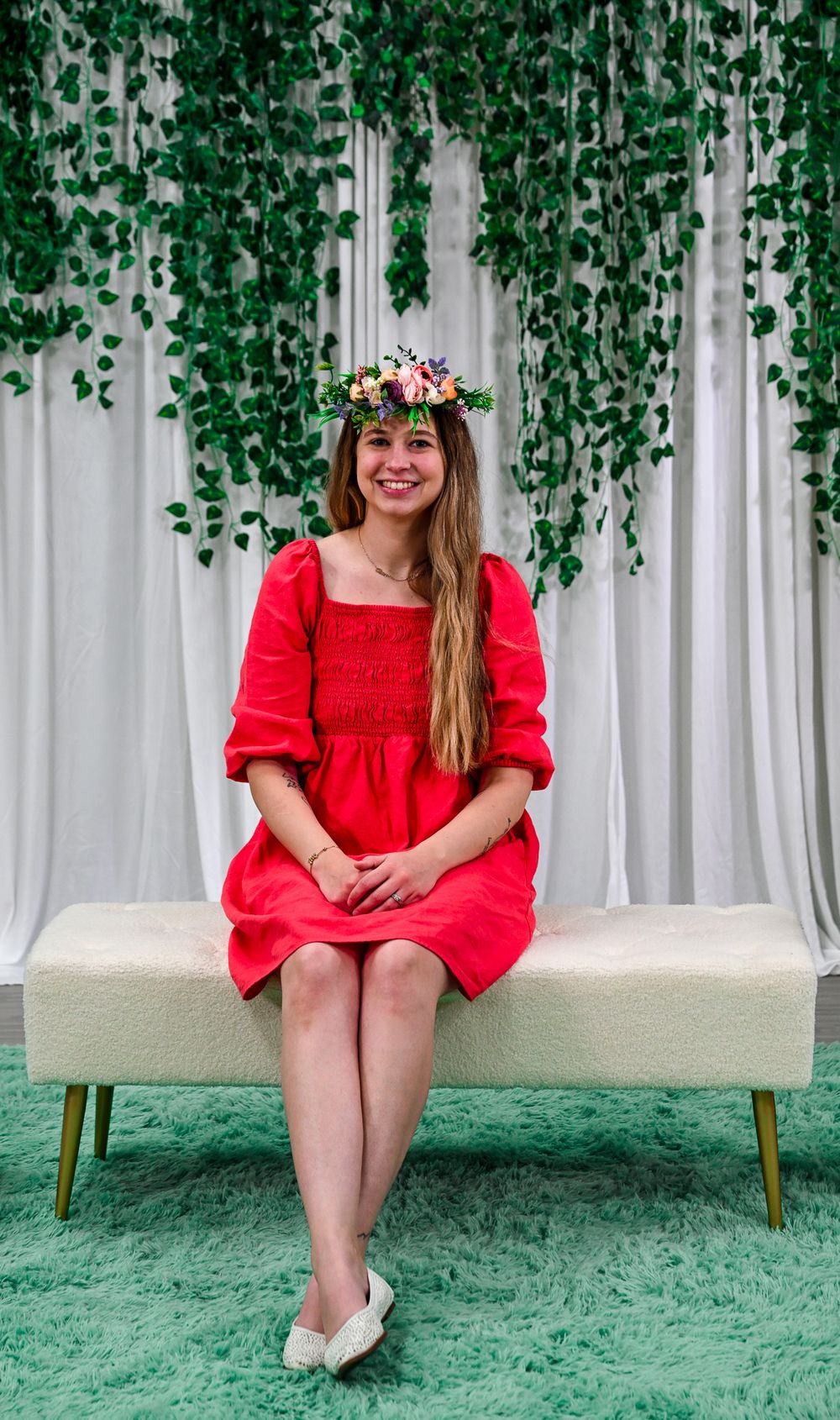 Smiling woman in red dress with floral crown sits on white bench against green ivy backdrop.
