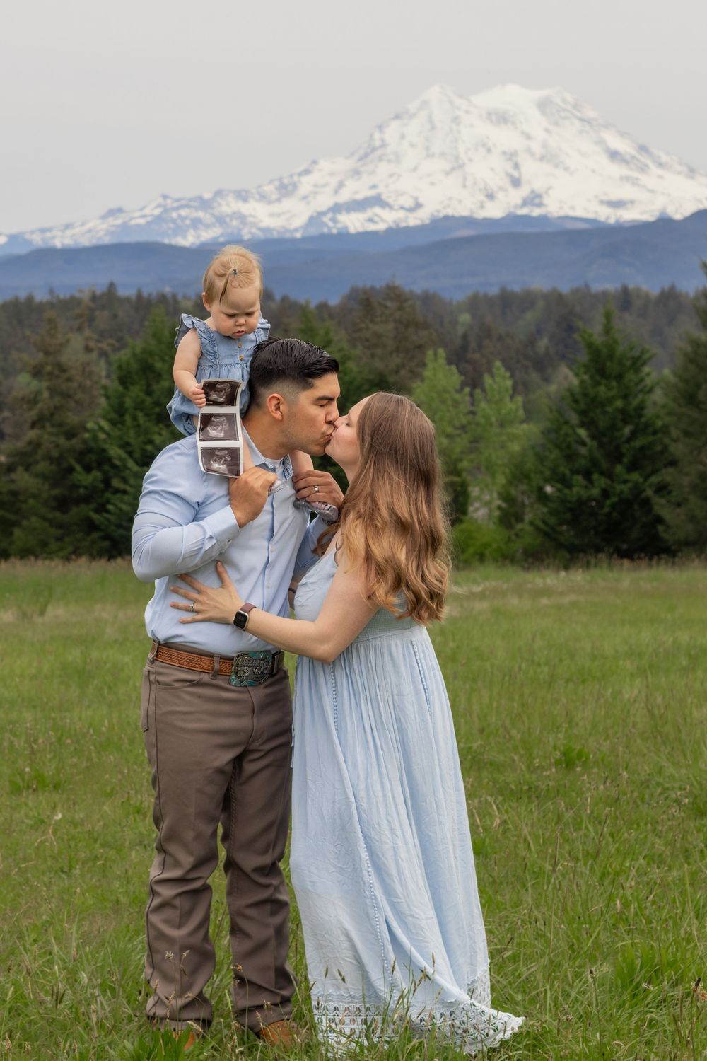 family of 3 in a green meadow with toddler holding ultrasound photos, snowy Mount Rainier in background. taken by roy wa photographer