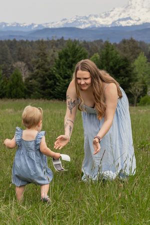 Mother in blue dress reaching for toddler holding ultrasound photo in a lush green meadow with mount rainier behind in eatonville wa
