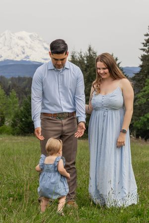 Parents in blue outfits watching their toddler walk in a green meadow with snow-capped Mount Rainier in background taken by Roy photographer
