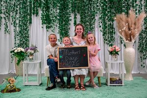A woman and three children hold a Mother's Day chalkboard sign in a decorated photo booth setting.
