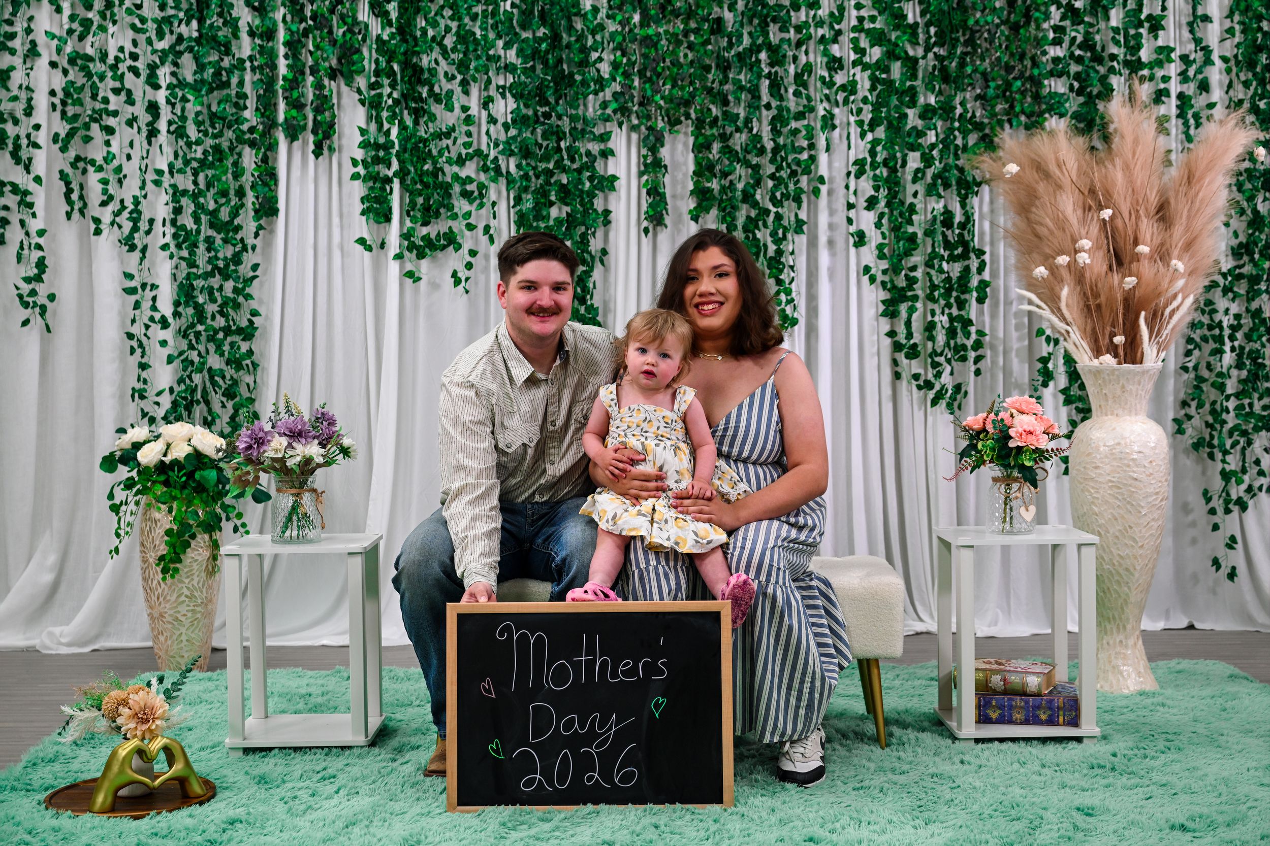 Family posing with a Mother's Day 2026 chalkboard sign in a decorated photo booth with greenery backdrop.