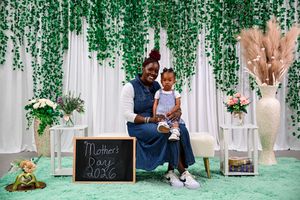 A mother and child pose together at a Mother's Day 2026 photo booth with green floral backdrop.