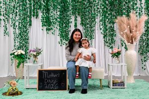 A mother and toddler pose together on a bench at a Mother's Day 2026 photo setup with green floral backdrop.