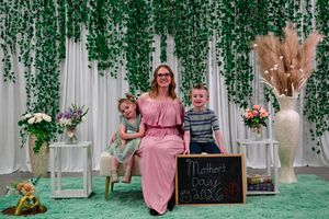 Woman in pink dress poses with two children for a Mother's Day photo in front of green ivy backdrop.