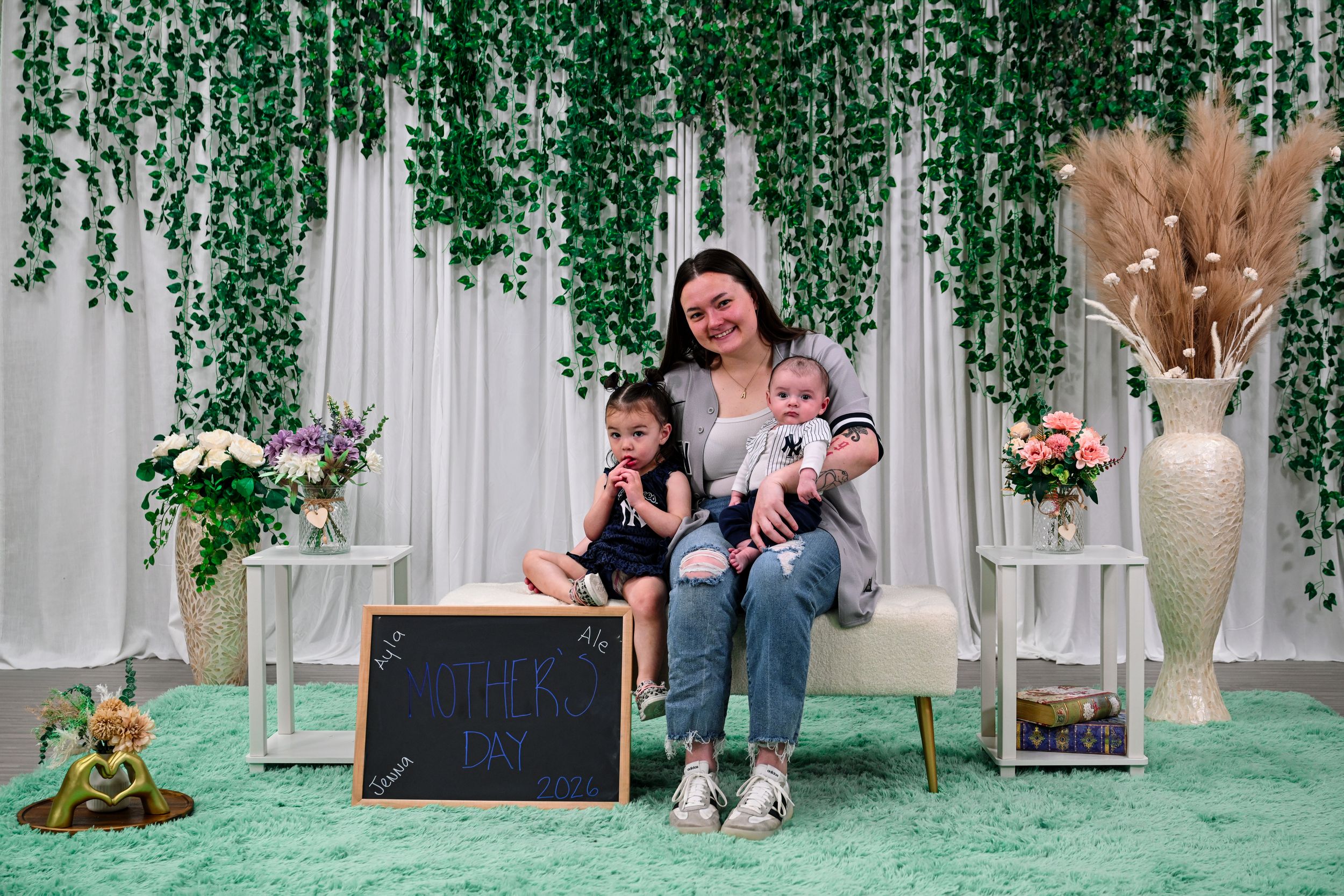 A mother sits on a bench with two young children in a decorated studio with green ivy backdrop for a Mother's Day photo.