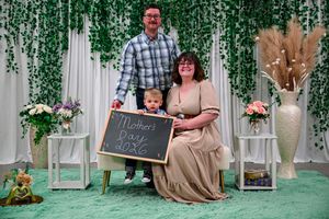 Family of three posing for a Mother's Day 2026 photo with chalkboard sign and green floral backdrop.