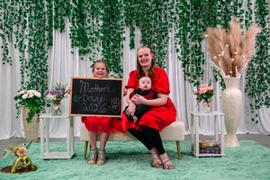 Woman in red dress holds baby while girl displays Mother's Day 2026 chalkboard sign in festive green backdrop.