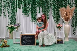 Mother and child pose together at a Mother's Day 2025 photo backdrop with green ivy, white drapes, and floral decor.