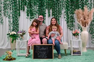 Family of four poses for Mother's Day 2026 photo in front of floral backdrop with green decor.