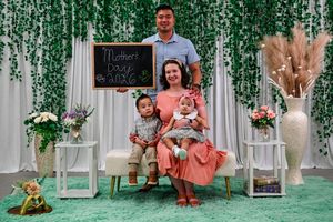 Family posing for a Mother's Day photo with a chalkboard sign, greenery backdrop, and two young children in a festive indoor setting.