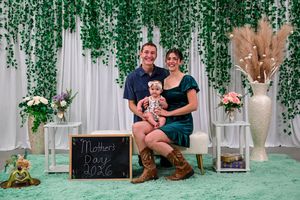 Young family with baby poses before green ivy backdrop with Mother's Day 2026 chalkboard sign.