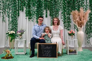 Family of three posing for a Mother's Day 2026 photo in front of a green ivy backdrop with floral decorations.