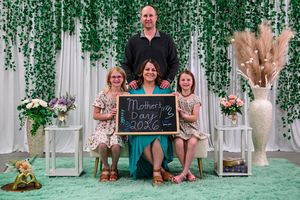 Family of four posing with a Mother's Day 2026 chalkboard sign in a decorated floral backdrop.