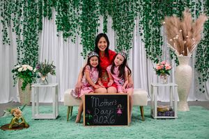 A mother and two daughters pose for a Mother's Day 2026 photo in matching pink outfits against a green floral backdrop.