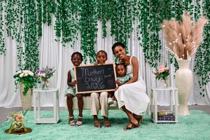 A mother and three children pose with a Mother's Day 2023 chalkboard sign against a green floral backdrop.