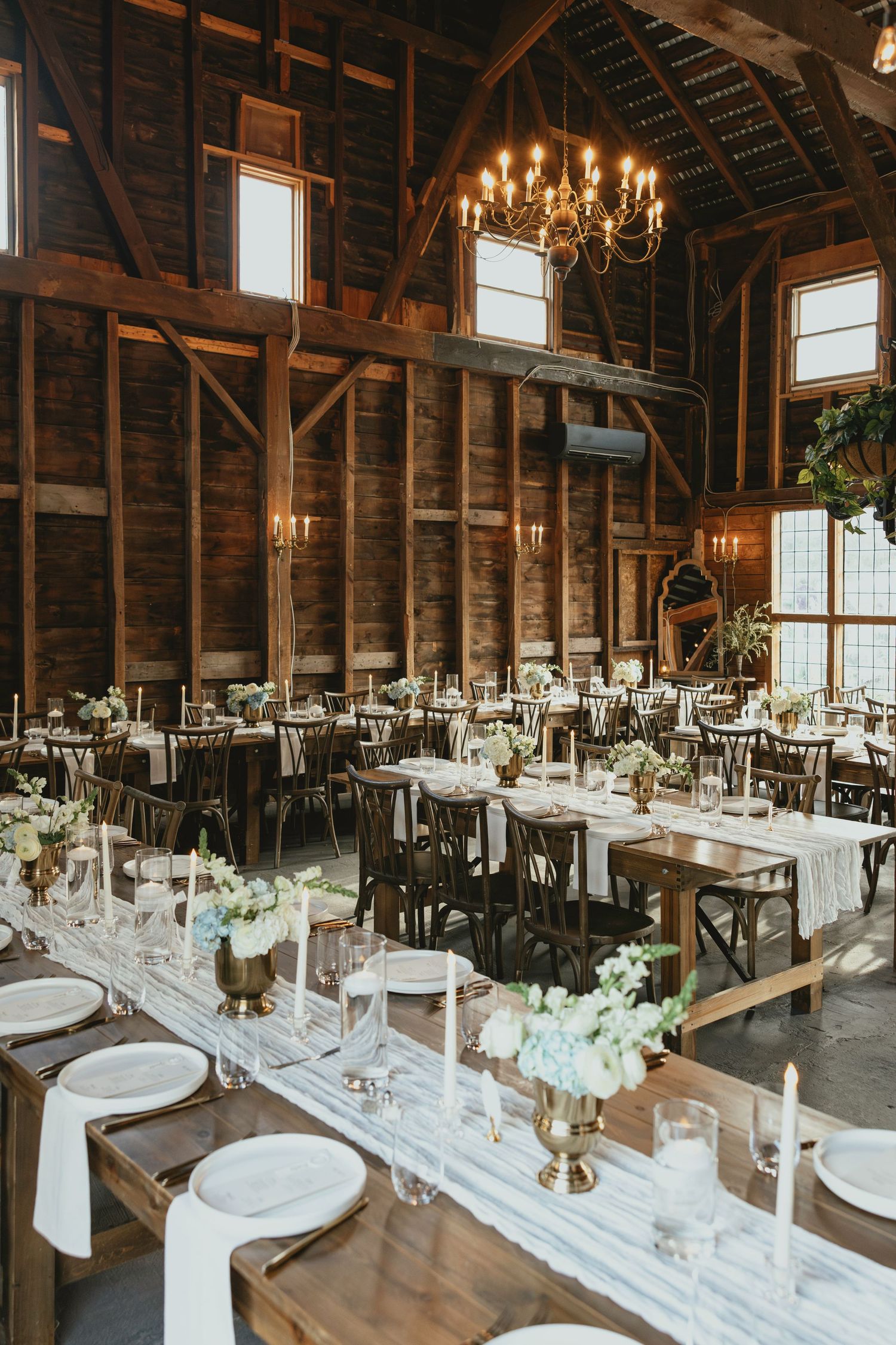 Rustic barn wedding reception with long wooden tables, white floral centerpieces, gold candelabras, and warm chandelier lighting.