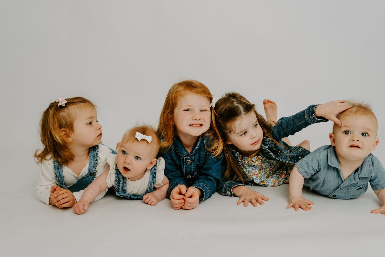 Five young children pose together lying down in a studio, smiling at the camera against a white background.