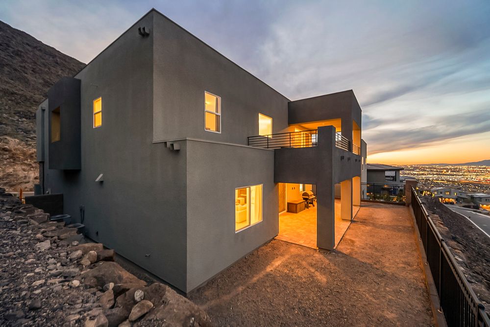 Contemporary gray stucco home on a hillside with city views and glowing interior lights at sunset.