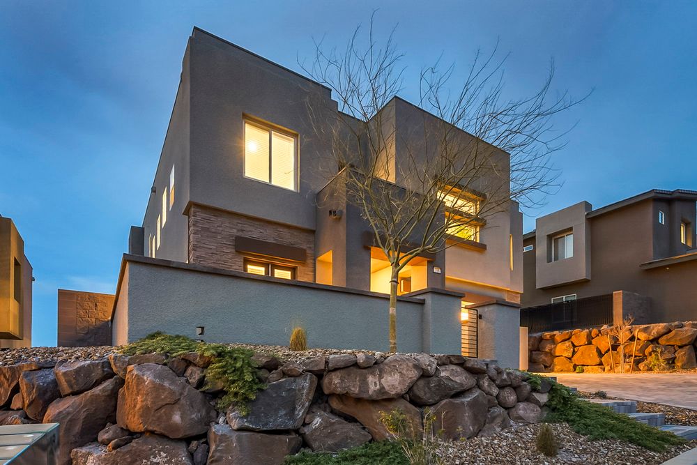Modern two-story stucco home with boulder landscaping and warm glowing windows at twilight.