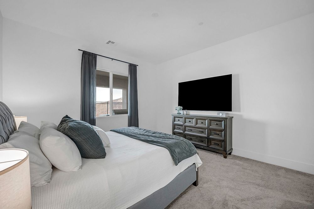 Modern bedroom with gray upholstered bed, plush carpet, large TV on dresser, and window with blue curtains.
