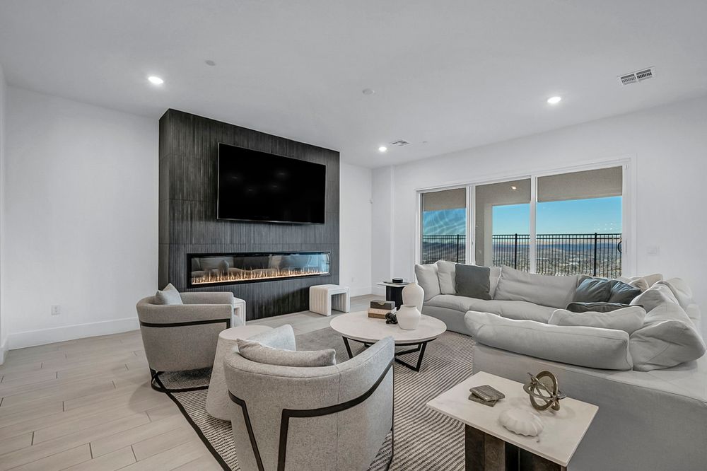 Bright living room with linear fireplace, wall-mounted TV, gray sofas, accent chairs, and panoramic windows.