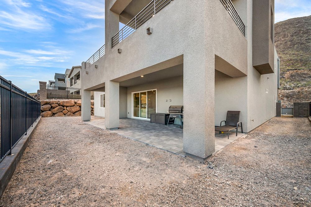 A modern two-story stucco home features a covered patio and gravel yard backing a rocky hillside.