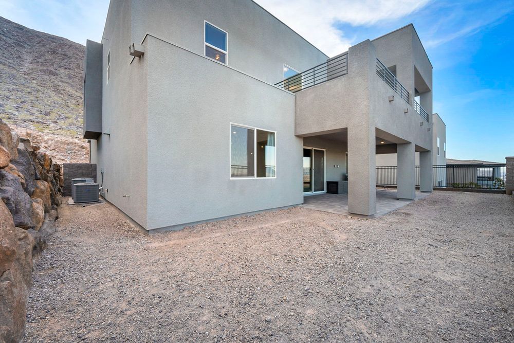 A contemporary two-story beige stucco house with a balcony sits against a rocky desert hillside.