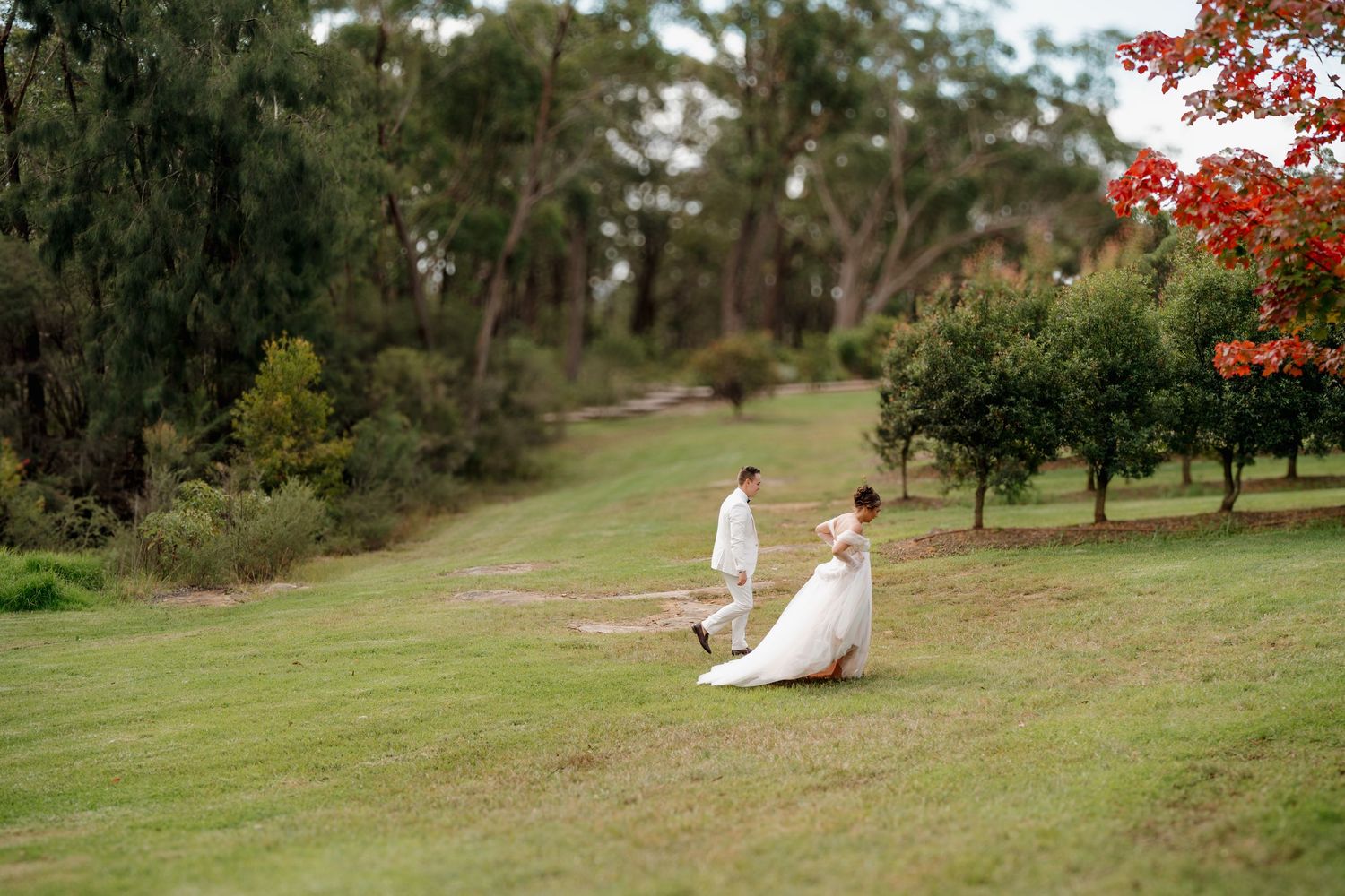 Bride and groom walking hand in hand across lush green lawn surrounded by trees at outdoor wedding venue.