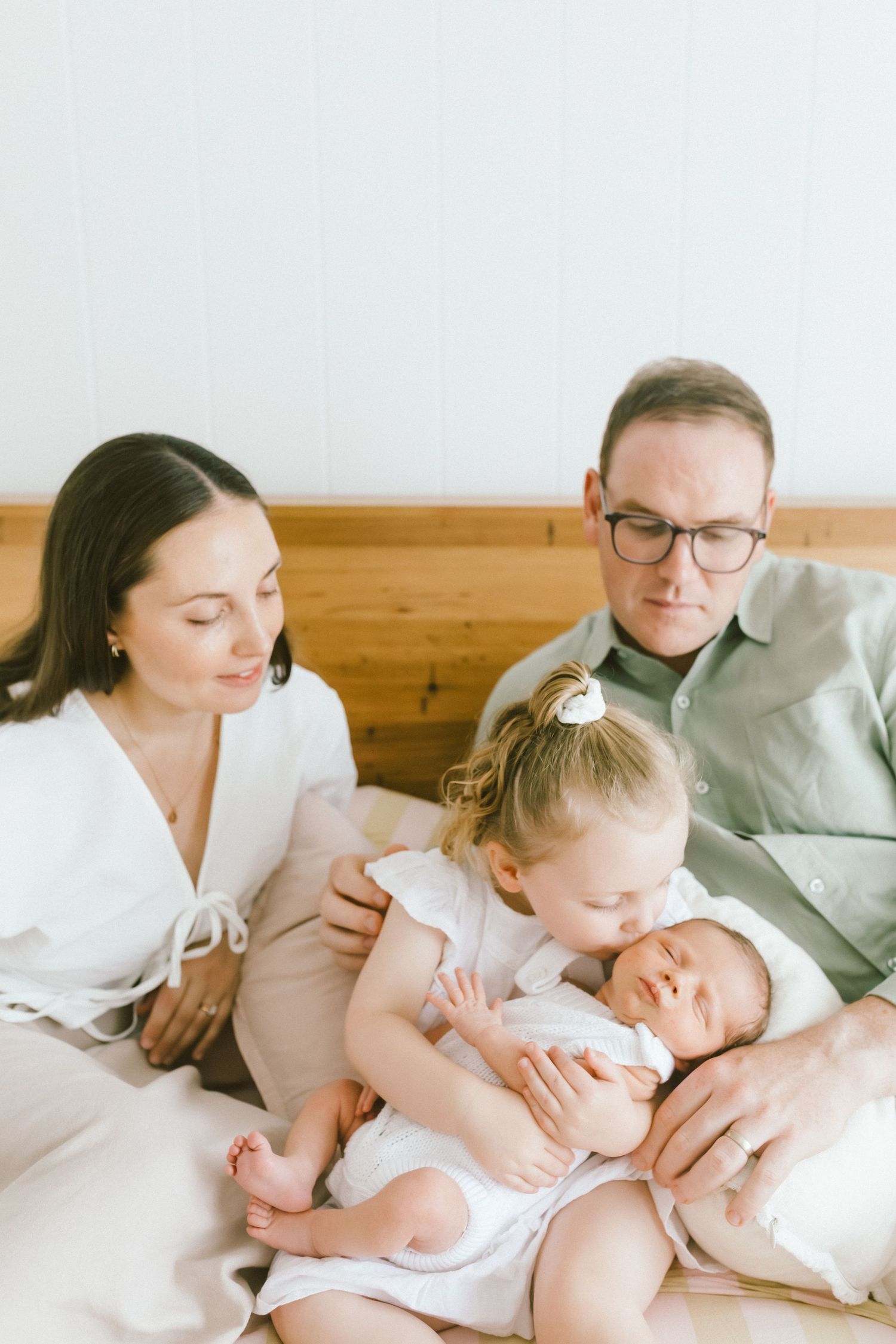A young family sits on a bed, parents gazing lovingly as their toddler kisses their newborn baby.