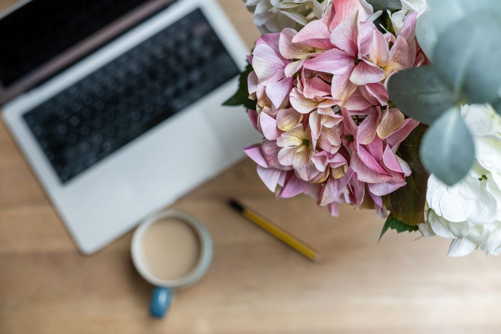 Overhead view of a workspace with pink hydrangeas, a laptop, coffee cup, and pencil on a wooden desk.