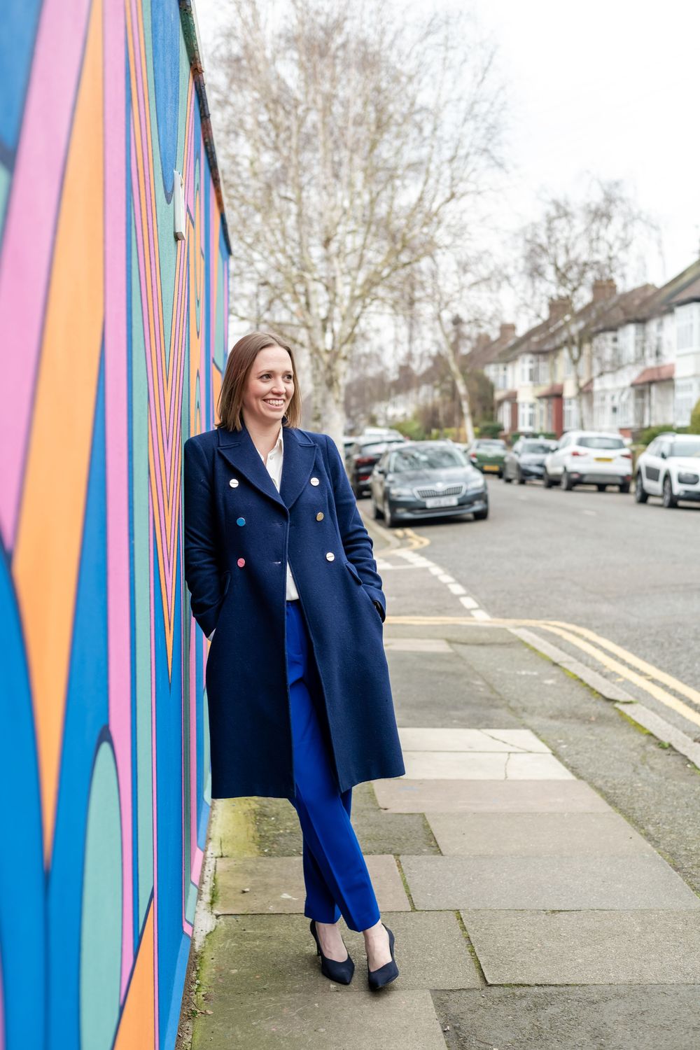 A smiling woman in a navy blue coat and trousers leans against a colorful mural wall on a residential street.