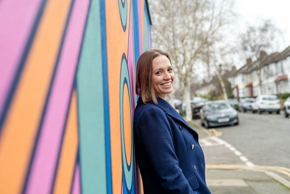 Smiling woman in navy coat leaning against vibrant colorful street mural in urban neighborhood.