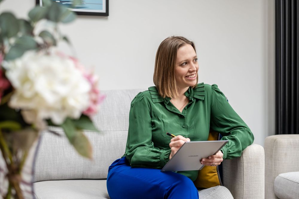 Woman in green blouse sitting on sofa with tablet, smiling in modern office setting.