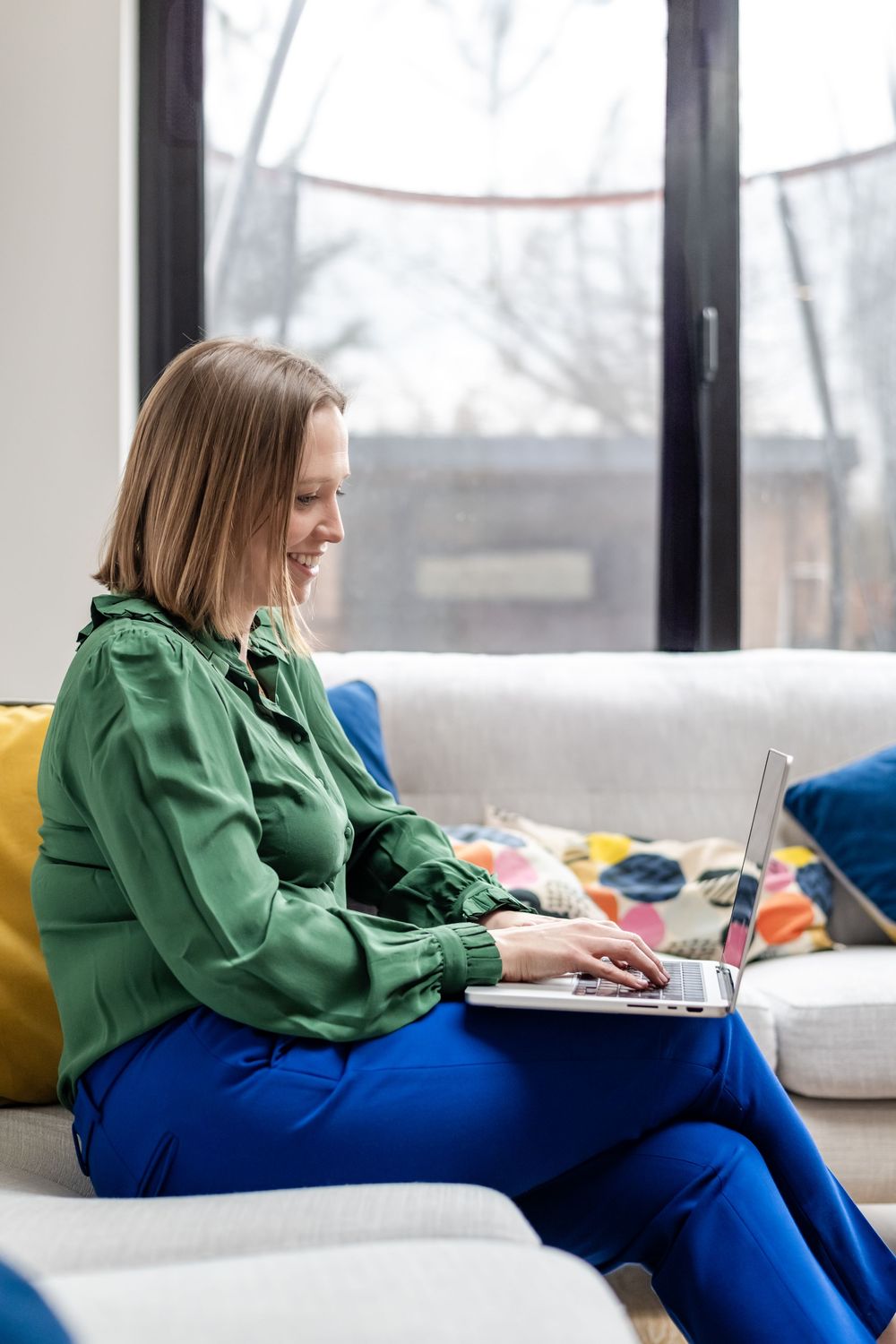 Woman in green blouse and blue trousers smiling while working on laptop seated on modern sofa.