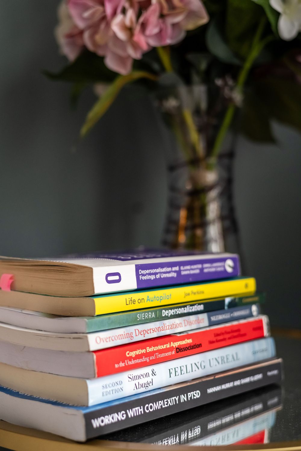 Stack of mental health books on depersonalization and PTSD beside a pink flower vase on a reflective table.