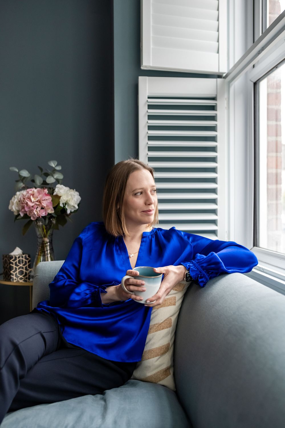 Woman in blue satin blouse sitting on sofa holding coffee cup, gazing out window with flowers nearby.