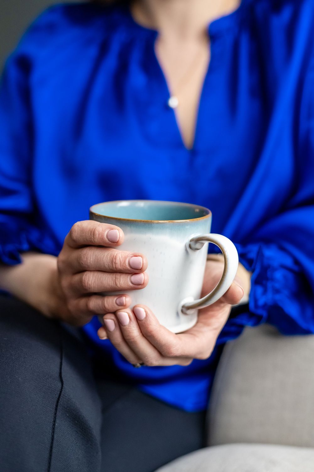 Woman in blue silk blouse holding a white ceramic mug with gold rim while seated on a gray chair.
