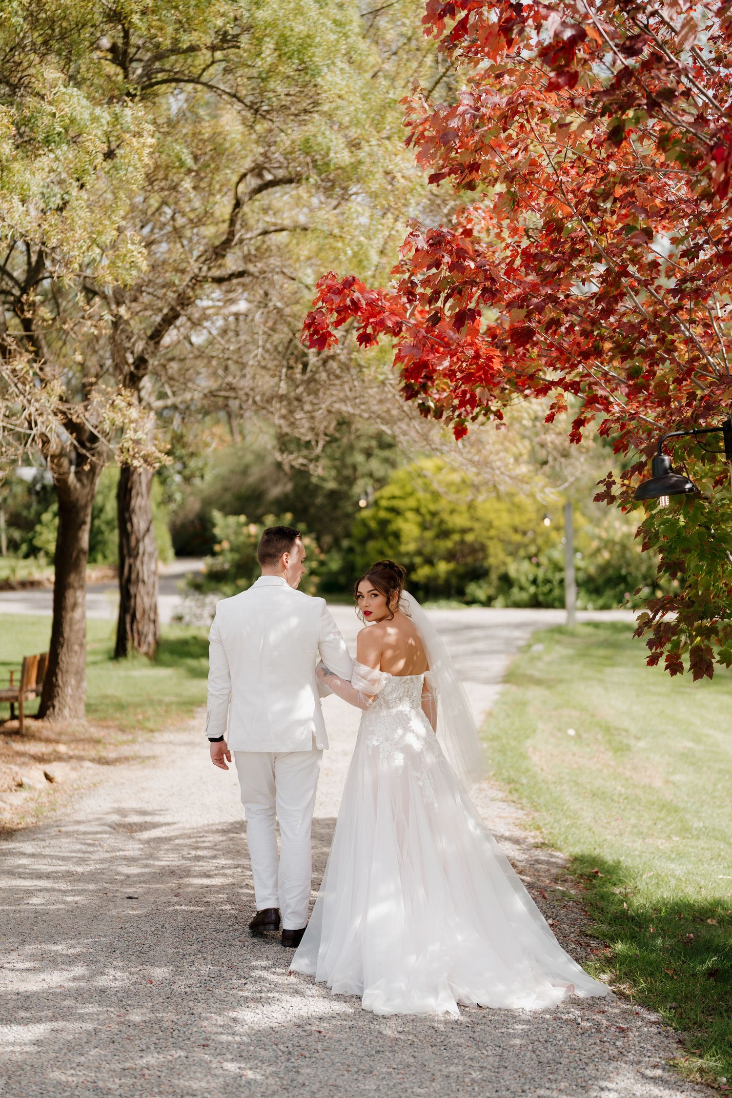 Bride and groom walking hand in hand on a garden path surrounded by lush green and vibrant red autumn trees.