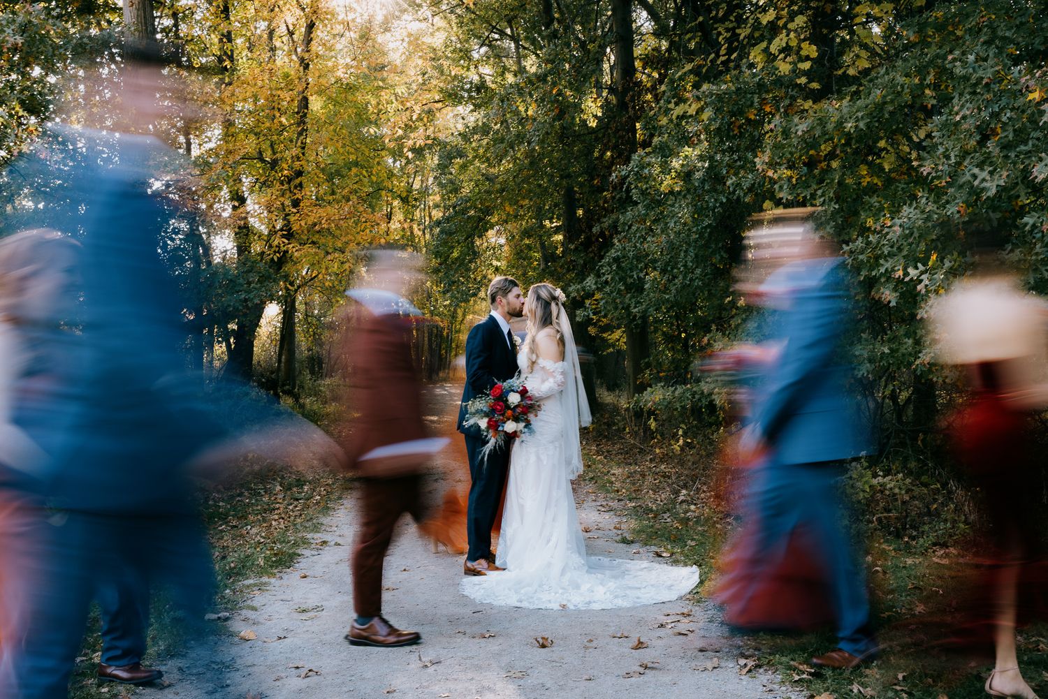 Long exposure of bridal party walking past bride and groom at Chagrin River Park in Cleveland in the fall with autumn foliage behind them.
