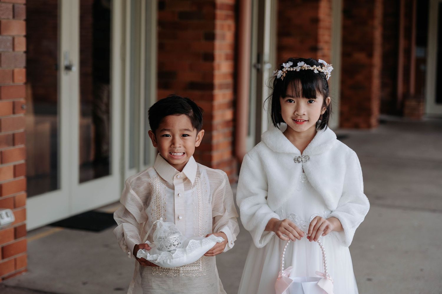 Two children dressed in elegant white outfits stand together in a formal celebration along a brick building walkway.