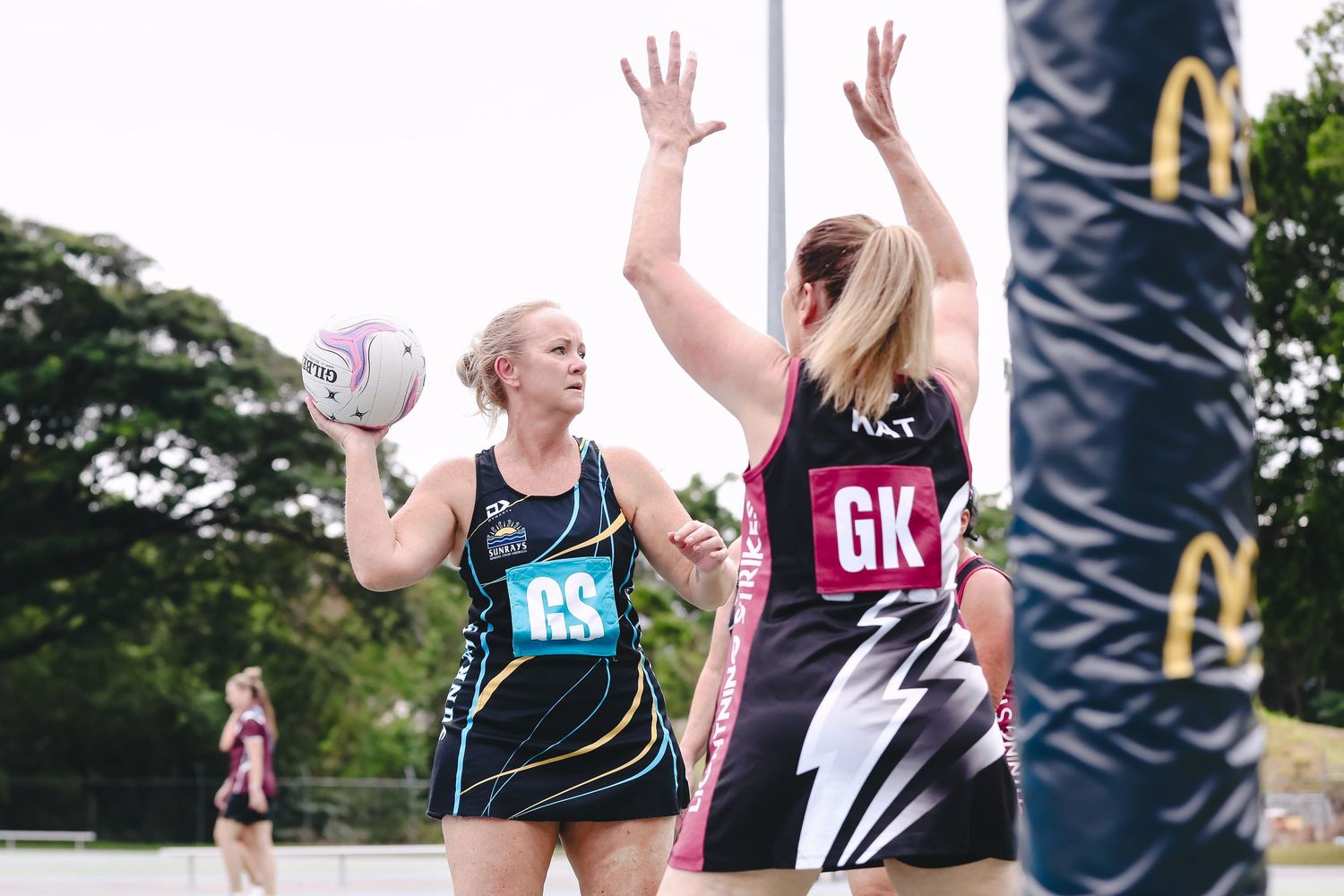Players competing in an outdoor netball match on a green court with trees in the background.