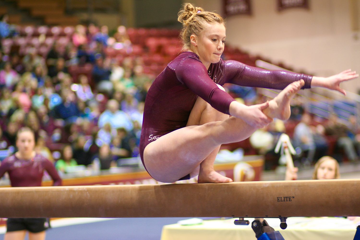Gymnast performing a precise balance move on the wooden beam during a competitive event in an indoor arena.