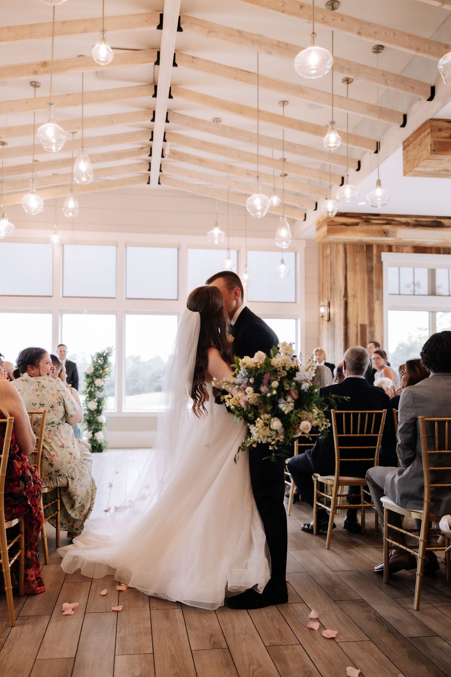 Wedding ceremony takes place outdoors under a floral arch with a couple exchanging rings near a waterfront setting.