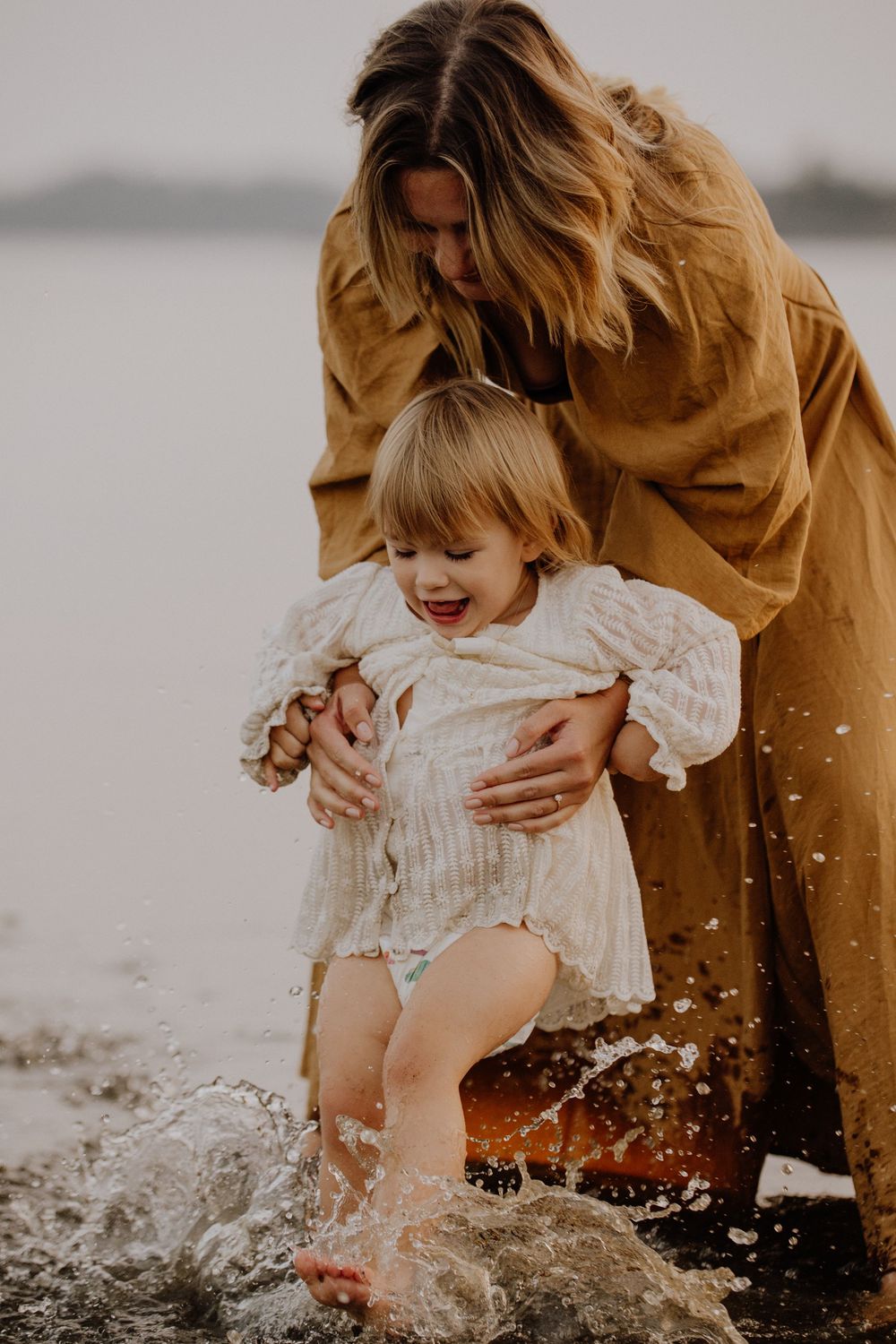 Parent and baby child splash joyfully in the water at the beach wearing warm brown and white clothing during sunset.