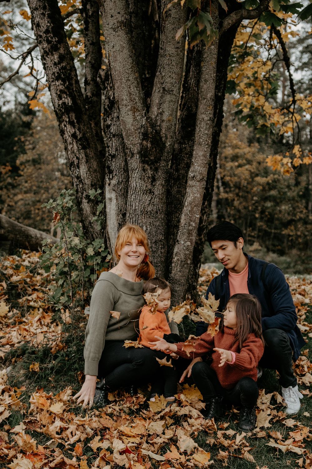 Family sits together surrounded by fallen autumn leaves beneath a large tree trunk on a moody fall day.